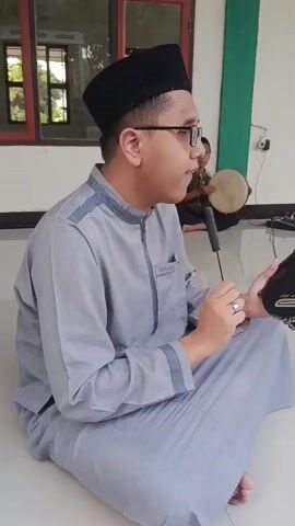 A man wearing glasses and a black cap sits cross-legged on the floor. He is dressed in a light grey long-sleeved shirt and matching pants. In his right hand, he holds a microphone attached to a stick, which he gestures with. His left hand rests on a black object with a reflective surface, possibly a drumhead. To his right, a black audio mixer with several knobs and buttons is visible. In the background, a young boy sits on the floor, also cross-legged, playing a small drum. The setting appears to be outdoors or in an open area with a tiled floor and a wall in the background.