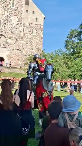 A person in full knight's armor, holding a shield, rides a gray horse adorned with a red and black tapestry. The horse is walking on a green grassy field. In the foreground, several people are seated at wooden tables, watching the spectacle. Some are holding up phones to record. Behind the knight and horse, a stone building with a tall tower and trees are visible. The sky is clear and blue, indicating a sunny day.