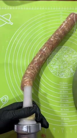 A close-up shot shows a pair of black gloved hands operating a sausage stuffer. The stuffer is filled with a mixture of ground meat and is connected to a casing that is being filled with the meat mixture. The sausage casing is long and coiled, and it is being fed onto a sausage stuffer attachment. The background is a green mat with circular markings and measurements. The lighting is bright and even.