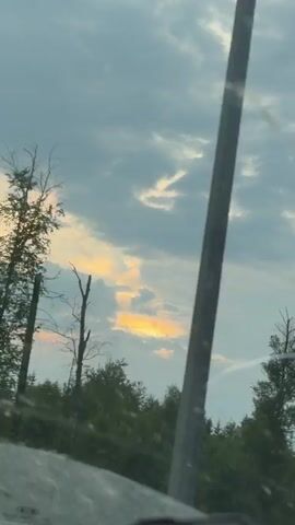 The video shows a time-lapse of clouds moving across the sky during what appears to be sunset. The sun is visible peeking through a gap in the clouds, casting a warm orange glow. Silhouetted against the sky is a line of trees, with some bare branches visible among the leafy ones. The lower left corner of the frame shows a portion of a vehicle, suggesting the footage was captured from inside a moving car. The overall impression is one of a serene natural landscape.