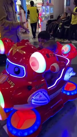 In a well-lit indoor amusement center, two young children are riding a bright red, coin-operated airplane ride. The older child, wearing a red shirt, is guiding the younger child, who is wearing a striped shirt, as they both grip the orange steering wheel. The ride features flashing colored lights and a small video screen that displays a virtual flying game. An adult in a light-colored shirt stands nearby, watching over the children. In the background, other arcade machines, people, and the center's decor are visible.