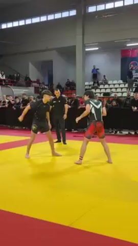 The video shows two people engaged in a stand-up grappling match on a mat. The mat has a yellow center and a red border. The grapplers are barefoot. One grappler wears black shorts and a rash guard with a design on the shorts. The other grappler wears black shorts and rash guard with a red pattern on the shorts and red on the top. A referee in black pants and a black shirt watches the grapple. A crowd watches the grapple from the stands.