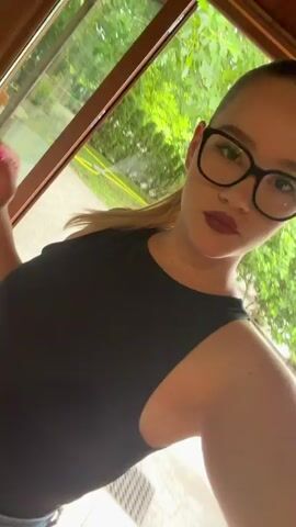 This media file features a young woman with blonde hair pulled back in a ponytail, wearing a black tank top and jeans. She has on large, black-rimmed glasses and dark red lipstick. Her fingernails are painted with bright pink and yellow polish.

The video appears to be a selfie taken indoors, with a window in the background showing a lush green outdoor environment, suggesting it's daytime with pleasant weather. The woman is posing for the camera, making kissing gestures and looking at the lens. Her facial expressions convey a playful and confident demeanor.

The interior setting shows hints of wooden furniture and tiled flooring. The overall ambiance is casual and relaxed. There is no visible text in the media file. The location context provided is Beguntsi, Bulgaria.