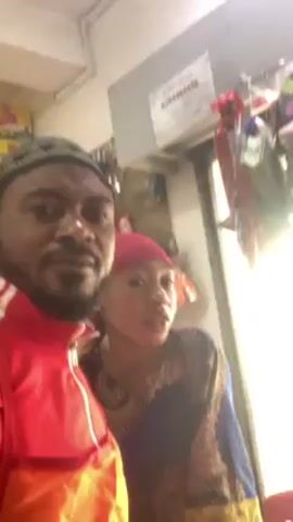 A man and a younger woman are standing close together, looking towards the camera. The man is wearing a red Adidas track jacket and a kufi hat. The woman is wearing a patterned top and a red headscarf. Behind them is a door and some shelves with various items. There is a sign with numbers on it above the door. The lighting is bright, potentially indoors. They appear to be posing for the video.