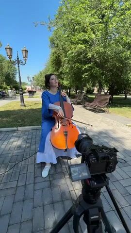 A woman is playing the cello in a park. She is wearing a blue dress and white shoes. The cello is orange. There is a camera on a tripod in front of her, recording her performance. The park has trees, benches, and lampposts. The sky is blue.