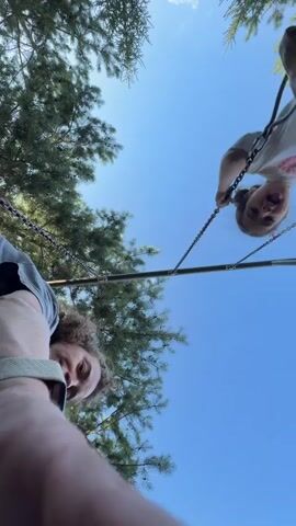 The video shows two young individuals on swings, viewed from below looking up at a bright blue sky. The perspective is upside down. On the left, one individual, with curly hair, wears a grey t-shirt and is holding onto the chains of a swing. On the right, another individual, wearing a white t-shirt, is also on a swing, holding the chains, with their head tilted. Both individuals appear to be in motion. Tall green pine trees frame the top of the shot, with their branches extending downwards. The swings appear to be made of dark material, possibly plastic, suspended by metal chains. The sun is shining, creating a bright and clear atmosphere.