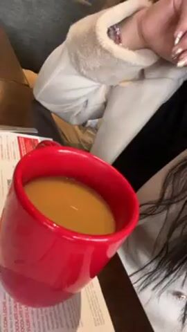 A woman with long dark hair sits at a table with a red mug of liquid in front of her. She wears a white jacket and a black turtleneck shirt. Her nails are painted with a gold and silver design. She looks at the camera and drinks from the mug. There is a menu on the table next to the mug. In the background, there is a person with curly blonde hair. The scene appears to be in a restaurant or cafe.