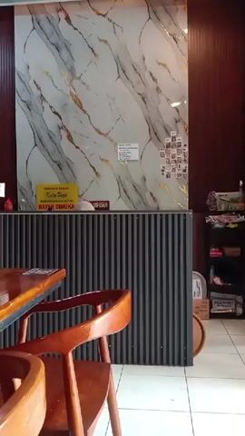 The video shows the interior of a restaurant. In the foreground are shiny wooden tables and chairs. In the background, a reception desk is visible with a marble-like wall behind it, adorned with photographs. The restaurant has a modern design with dark red and black accent walls. Two women are sitting at a table in the background, watching television. The interior appears clean and orderly.