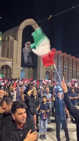 The video shows a nighttime outdoor crowd of people gathered in front of a large building with an arched entryway. A large banner with a portrait of a man in religious attire is displayed in the arch. Many people in the crowd are holding up Iranian flags and other banners, some of which are red. The crowd appears to be moving or gathering. Some individuals are seen raising their hands and waving. The atmosphere seems to be one of a rally or protest. The lighting suggests it is dark, and the event is taking place at night.