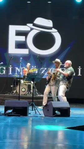 A man in a straw hat and a light-colored shirt and pants plays an electric guitar and sings on a stage. He is standing in front of a music stand with sheet music. To his right, another man with a white beard and a patterned shirt plays a saxophone. Behind them, a drummer is partially visible, playing a drum set. The stage has black speakers in the foreground and a large screen behind the musicians displaying the letters "EO" with a fedora hat superimposed over the "O". The lighting on the stage is blue and white. The video appears to be a live performance.