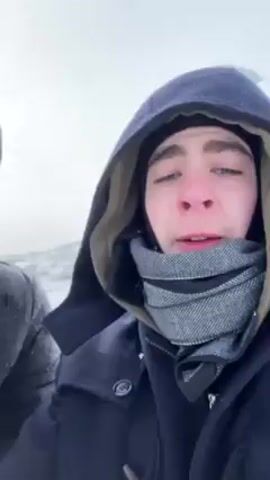 Two young men, bundled in winter clothing, stand in a snowy landscape, holding cans of Bohs beer. The man on the right wears a hooded navy coat and a gray scarf. The man on the left is in a dark hooded jacket and black gloves. The background is a white, snow-covered terrain. The sky is overcast.