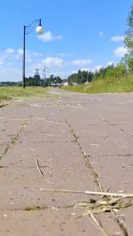A young child walks away from the camera on a paved path under a bright blue sky with scattered clouds. The child is wearing a light-colored onesie with a print and white shoes. Green grass and trees are visible on either side of the path, and a lamppost is in the background. Some dried grass and debris are on the paved path in the foreground.