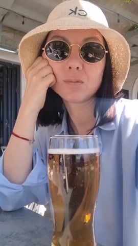 A woman wearing a light blue shirt, round sunglasses, and a straw hat with the letters "CK" on it is sitting at an outdoor table. She has short black hair and is resting her head on her left hand. In her right hand, she holds a tall glass filled with amber-colored beer. She takes a sip of the beer, holding the glass up to her mouth. The background shows a building with gray stone and black-framed glass doors. Sunlight is visible, creating highlights on the objects and person in the frame.