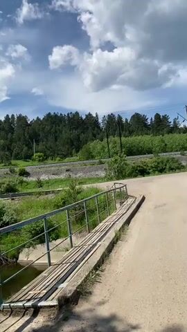 The video begins with a view of a dirt road leading to a wooden bridge over a body of water. A metal railing lines the side of the bridge. Beyond the bridge, there is lush green vegetation and a dense forest. In the background, a train track runs through the landscape, and further back, the forest continues to cover rolling hills. The sky is a clear blue with scattered white clouds. The camera pans across the scene, showing the river and the surrounding greenery. The video ends with a wide shot of the river flowing through a wooded area.