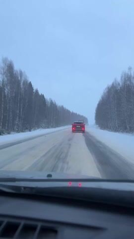 The video shows a car's perspective driving on a snow-covered road. A red car is visible ahead, and trees line both sides of the road. The sky is overcast and grey. The windshield wipers are not activated. Red lights are reflected on the dashboard of the filming car. The road surface appears wet or icy.