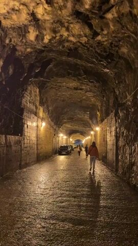 The video shows an underground tunnel. The tunnel is constructed of natural rock and is dimly lit with yellow lights. The floor is wet and shiny. Several people are walking away from the camera toward the end of the tunnel. Cars are parked on the left-hand side of the tunnel.
