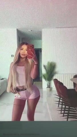 A woman with long blonde hair, wearing a pink crop top and pink shorts, stands in a room and takes a selfie with her phone. She is looking at the camera and holding her phone in her right hand. There is a plant in the background and a chair. The room appears to be a modern apartment.