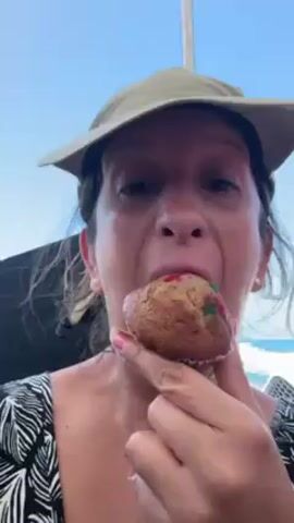 A woman wearing a khaki visor and a black and white patterned top eats a muffin with candied fruit pieces. She takes a bite, chews, and looks directly at the camera. The muffin is in a clear, plastic wrapper. In the background is a body of water and a white structure. A man in a light blue shirt can be seen to the right of the woman.