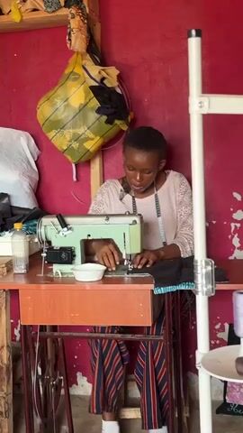 The video shows a young woman sitting at a sewing machine and working on a piece of dark fabric. She's wearing a light-colored, lacy long-sleeve shirt and striped pants, with a measuring tape around her neck. She is focused on her work, using both hands to guide the fabric under the needle of the vintage green sewing machine.

The sewing machine is sitting on a wooden table. A small white bowl sits near the machine. Behind her is a red wall on which hangs a yellow plastic bag filled with various fabrics. There is a white garment bag leaned against the wall to the left of the woman, and a white clothing rack is visible on the right side of the frame. There is a bottle of water on the table beside the sewing machine.