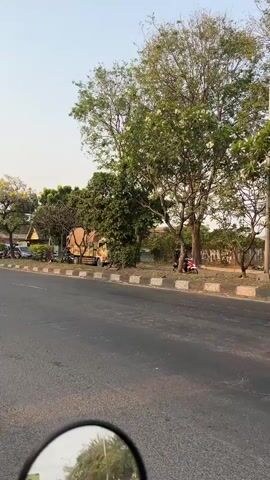The video shows a street scene with several vehicles and people. A yellow truck with a tan tarp covering its cargo is parked on the right side of the road. The truck has writing on its side, including "DIESEL" and "AMARAH". Two motorcycles are visible. On the left side of the road, a red motorcycle is parked, and a person is sitting on it. Further down the road on the right side, a red motorcycle is being ridden by a person wearing a helmet. Trees and foliage are present, with some branches obscuring parts of the vehicles. The road has a concrete curb and a sidewalk. The overall environment appears to be outdoors, possibly in a town or city.