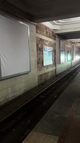 A subway train is approaching a station platform. The train is illuminated by its headlights, which are shining brightly. The station platform is made of stone and has advertisements on the walls. The train tracks are wet, suggesting that it may have recently rained or that the tracks are being cleaned. The lighting in the station is dim, with the exception of the train's headlights and the illuminated advertisements. The station appears to be empty of people, with the exception of the approaching train.