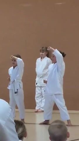 In a room with a brown wall, three young girls in white karate uniforms stand on a wooden floor. The girl on the left, wearing a white belt, has her left hand raised to her forehead and her right fist held near her hip. The girl in the middle, also wearing a white belt, stands with her arms at her sides. The girl on the right, with a white belt, has her right arm raised with her fist in the air and her left fist near her hip. The girl on the right appears to be in motion, and she is looking forward. The other two girls are also looking forward. The video seems to capture a moment during a karate lesson or demonstration.