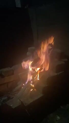 The video shows a campfire burning at night. The fire is contained within a makeshift enclosure of bricks. Flames, ranging in color from orange and yellow to purple, are visible dancing above the burning logs. The surrounding environment is dark, suggesting it is nighttime. The camera is positioned at eye level, facing the fire. There are no people or other objects clearly visible in the frame besides the fire and the bricks.