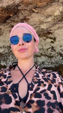 A woman wearing a pink headscarf and blue reflective sunglasses stands on a sandy beach in front of a rock face. She wears a black swimsuit top with crossed straps and a pink and brown leopard print cover-up. She is speaking and appears to be filming herself. The camera pans up to reveal a blue sky and green foliage above the rock face. The camera pans again to show people relaxing on blankets on the beach.