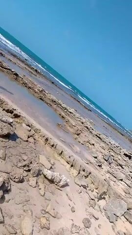 The video shows a rocky beach landscape. Initially, the scene is a static shot of the beach, revealing a mix of sand, rocks, and small pools of water under a clear blue sky. The ocean is visible in the background. Subsequently, a person wearing a blue long-sleeved shirt and dark shorts walks into the frame. They are seen walking barefoot across the rocky terrain near the water's edge. Another person is briefly visible, walking near the first person. As the video progresses, the camera angle changes, displaying a wider view of the beach and ocean, now under a sky with some cloud cover. The overall environment suggests a coastal area with a rocky shoreline.