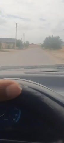 The video shows the interior of a car as it drives down a residential street. The camera is positioned to capture the road ahead and the driver's hand on the steering wheel. The street is lined with houses and fences, and there are power lines overhead. The sky is cloudy. The car appears to be moving at a slow to moderate speed.