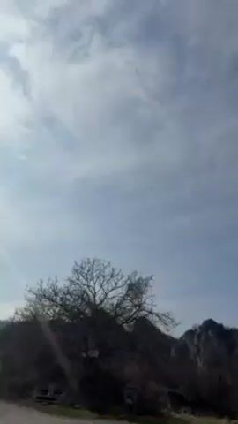 The video shows a landscape of rocky hills against a blue sky with cirrus clouds. The foreground includes a bare tree and some greenery, suggesting early spring. The camera pans slowly to the right, revealing more of the hilly terrain. The overall impression is one of a rural, possibly mountainous area with a clear sky.