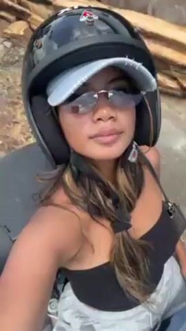 A young woman is riding a motor scooter. She is wearing a black helmet, a blue denim cap, and mirrored sunglasses. She has long brown hair with blonde highlights. She is wearing a black tank top and grey shorts. The scooter has a black storage compartment on the back. The woman is taking a selfie video while riding. The background shows a street with buildings on either side. The sky is cloudy. The scooter appears to be moving at a moderate speed.