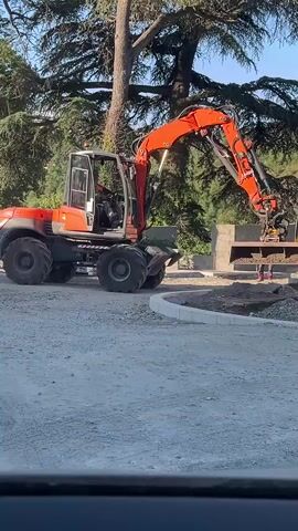 An orange excavator is positioned on a gravel surface. The excavator's arm is extended, and its bucket is lowered towards a concrete structure. A person in orange work attire stands near the concrete structure. Behind the excavator, a large tree with dense foliage is visible. In the background, there are more trees and what appears to be a building. The scene depicts a construction or demolition site.