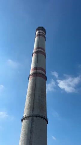 The video shows a tall, cylindrical chimney against a bright blue sky with scattered white clouds. The chimney is made of concrete and has red and white bands at intervals along its height. The camera angle is looking upwards from the base of the chimney, giving a sense of its immense scale. The lower portion of the video reveals the top edge of a large, dark, circular structure, possibly the opening of another part of the industrial facility, and a glimpse of the landscape below, which appears to be flat and somewhat arid. There are no people visible in the video. The overall impression is one of industrial infrastructure in an open, possibly rural, environment.