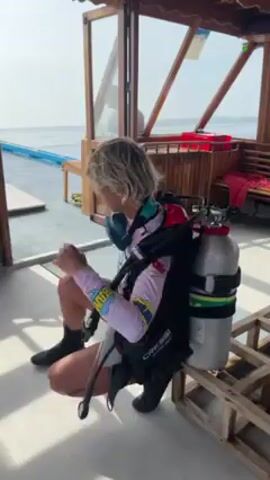 A woman with short blond hair is getting ready for a scuba dive inside what looks like a boat's cabin. She is wearing a light pink long-sleeved shirt with colorful graphics, black shorts, and black neoprene booties. She has a black buoyancy compensator device (BCD) and two scuba tanks strapped to her back. The tanks are silver, and one has a green band around it.

She starts in a squatting position, adjusting the BCD straps. Then, she stands up, further adjusting the straps and equipment on her chest. At one point, she picks up a piece of paper or laminated card, possibly a checklist, and looks at it.

The cabin has a wooden floor and ceiling. Outside the cabin windows, the ocean is visible under a sunny sky. Other objects in the cabin include a wooden bench with life jackets, a small table, and some pairs of green and white sandals. The sounds of the boat engine are audible.