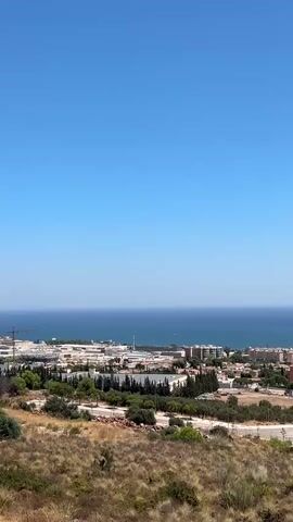 The video shows a wide shot of a coastal town under a clear blue sky. In the foreground, dry, scrubby vegetation covers a hillside. Below the hill, a mid-sized town with predominantly terracotta-colored buildings spreads out towards the ocean. The buildings appear to be apartment blocks and some commercial structures, with some trees interspersed. The ocean is visible beyond the town, stretching to the horizon. A few distant boats can be seen on the water. The scene is static, with no visible people or activity. The overall impression is a sunny day in a Mediterranean coastal area.