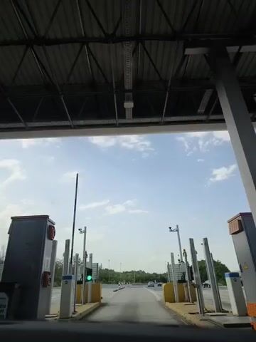 The video shows a view from inside a vehicle approaching a toll booth. The perspective is looking forward through the windshield. The toll booth structure is visible overhead, supported by pillars. On either side of the road leading to the toll booth are electronic payment machines. A green traffic light is illuminated on the left side, indicating passage is permitted. The road ahead is mostly empty, with a few vehicles visible in the distance. The sky is partly cloudy and bright.