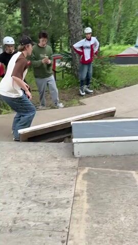 Two young people are in a skate park in a wooded area. The person in the foreground is wearing a brown and tan t-shirt, blue jeans, and a black baseball cap. This person is on a skateboard and is performing a grind on a red metal rail. The person in the background is wearing a red and white jacket, a white helmet, and blue jeans. This person is watching the skateboarder. The skate park includes ramps and rails. There are trees and green grass surrounding the skate park.