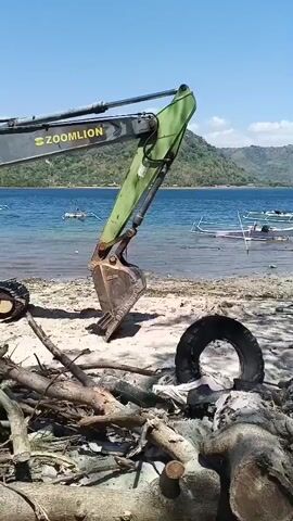 An excavator is shown on a sandy beach, with its bucket extended towards the water. The excavator is green and gray and has the word "ZOOMILION" on its side. There are piles of driftwood and debris on the beach, as well as a deflated tire. In the background, there is a body of blue water with several boats floating on it. Further in the distance, there are lush green hills under a clear blue sky. The camera pans slightly to the right, showing more of the beach and the surrounding environment.