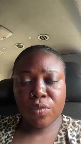 A close-up video shows a young woman with dark skin and braided hair looking directly at the camera. She is wearing a leopard-print top and has a nose ring and earrings. The background appears to be the interior of a vehicle, with air vents and a ceiling visible. The woman blinks and her expression changes from neutral to a slight frown as she looks downwards.