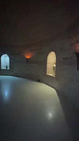 The video shows a slow panning shot of a circular, dimly lit interior space with brick walls and arched openings. The floor appears to be smooth and reflective. In one of the arched openings, there is a bright light source illuminating a pile of rocks. The ceiling is also visible and seems to be domed. The overall impression is of an ancient or underground structure. There are no people or other objects visible in the scene besides the structural elements and the lighting.