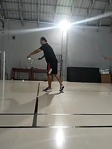 The video shows two people playing badminton indoors. The court is made of polished linoleum with black lines marking the boundaries. The ceiling is high and covered with corrugated metal panels, with bright overhead lights illuminating the court. A net divides the court in half. In the foreground, a person in a black shirt and red-and-black shorts is in the process of hitting a shuttlecock with a badminton racket. In the background, another person, wearing a lime green and white shirt and black shorts, is positioned on the other side of the net. There are also some tables and chairs visible in the background, as well as some bags and other sports equipment. The overall impression is that of a casual game of badminton being played in a community sports hall.