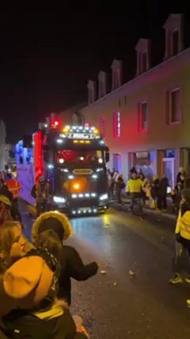 The video shows a nighttime street scene, possibly a parade or celebration. A large black Scania truck, brightly lit with many lights, is moving slowly along the street, surrounded by crowds of people. Some people are throwing objects, perhaps candy, into the crowd. The buildings on either side of the street are illuminated with festive colors, like red and white. People are standing on the sidewalks and in the street, watching the truck pass. Many are wearing reflective vests, and the general atmosphere is lively and festive.