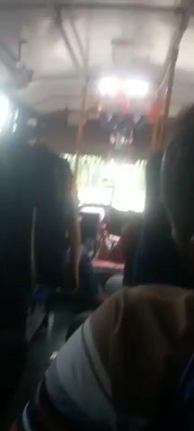 The video shows the interior of a moving bus. Several passengers are visible, some with their backs to the camera. The lighting appears to be natural, coming from the windows. In the foreground, a person's shoulder is visible, covered by a light-colored garment. In the middle ground, a person with dark hair is wearing a blue and white striped shirt and appears to be leaning back. Further down the aisle, another passenger is visible in a dark shirt and jeans. The overall impression is that of a typical bus journey.