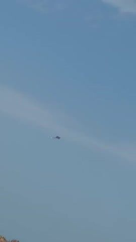 A kite with a purple and white design is seen flying against a pale blue sky with a wispy white cloud. The kite is positioned in the upper right quadrant of the frame. The bottom portion of the video shows a blurred, brown thatched roof-like structure, suggesting an outdoor setting.