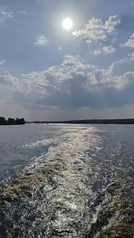 The video shows a wake behind a boat moving across the water under a bright, sunlit sky dotted with puffy clouds. The sun reflects brightly on the water's surface, creating a shimmering effect. The wake is prominent, a foamy trail of disturbed water directly behind the boat, diminishing into the distance. The water is relatively calm, and land can be seen faintly on both horizons.