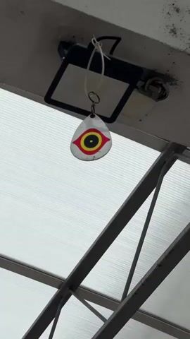 The video begins with a close-up of a shiny, tear-drop shaped object with a target-like design, hanging from the ceiling, beneath a square spotlight. The camera then pans out to reveal a covered walkway, constructed with metal beams and a translucent roof. A red carpet runs along the walkway, with benches on the right side. People can be seen walking along the walkway, and buses are parked on the left. There are also signs visible, and air conditioning units are mounted on the walls.