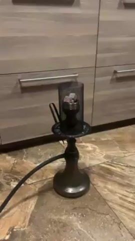 The video shows a hookah on a tiled floor. The hookah is gray and black, and has a long, black hose. The hookah bowl has a metal screen cage around it, with red-hot coals inside. There are kitchen cabinets in the background.