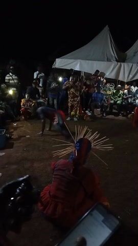 At night, a crowd surrounds a performance area on a dirt surface. Under a white tent, people sit on folding chairs. A man in red pants and a patterned blue shirt does a flip. Another person in red with wooden sticks jutting from their head sits on the ground in the foreground. A hand holds a lit cell phone.