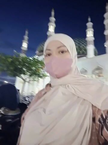 The video begins with a close-up of a woman wearing a light pink mask and a beige hijab and scarf. In the background, a mosque with a green dome and tall minarets is visible, illuminated against a dark blue twilight sky. As the video progresses, the camera pans slightly to the left, revealing a young girl in a beige hijab standing next to the woman. The girl is holding some items in her hands. Several motorcycles are parked in the foreground, and other people and vehicles are visible in the distance. The scene appears to be an outdoor area, possibly a parking lot, adjacent to the mosque. The overall lighting suggests it is evening or night.